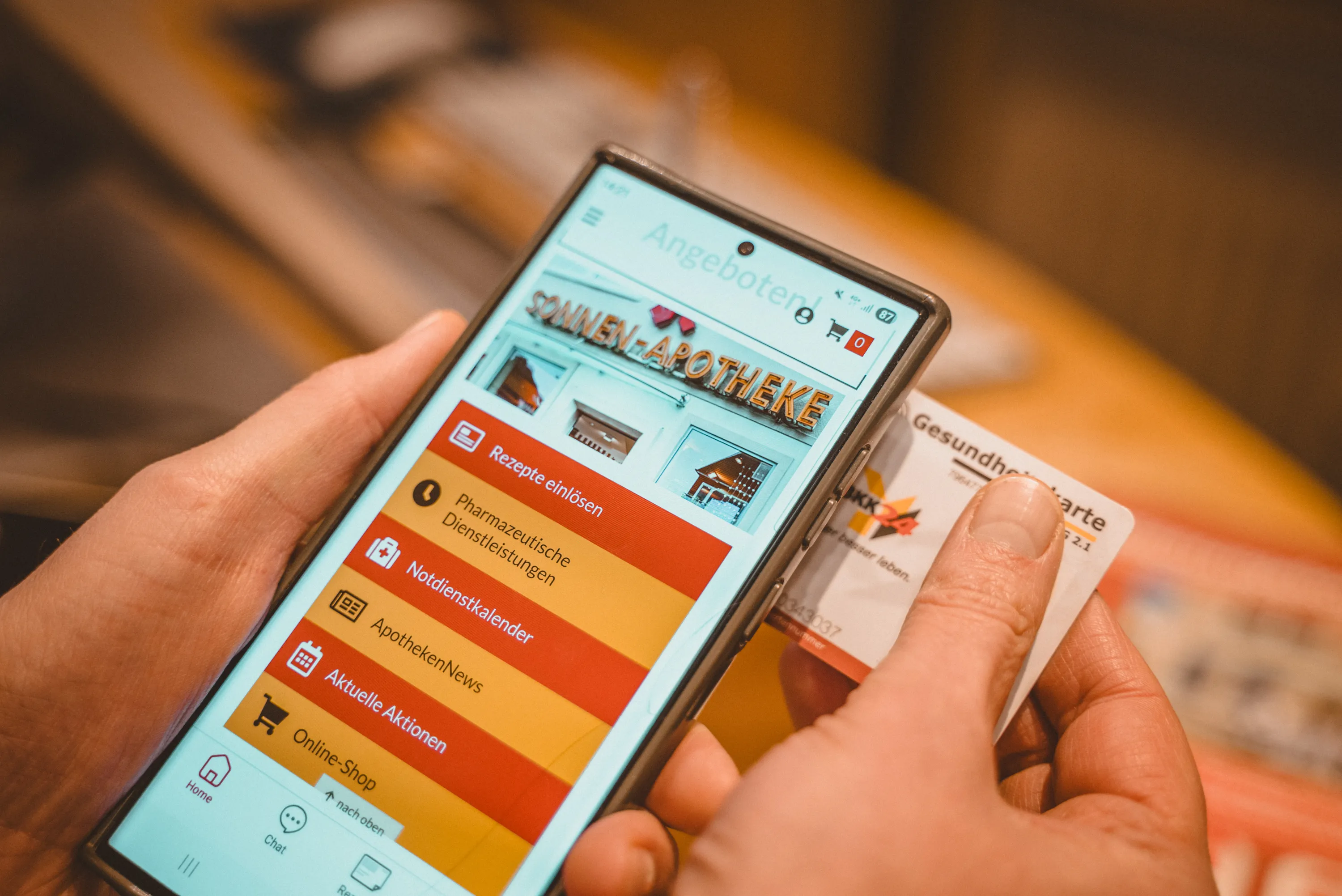 Hände halten Smartphone mit Sonnen-Apotheke-App und Krankenversichertenkarte